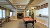 This single-floor amenity space is thoughtfully designed to serve as a premier entertainment and social hub. The standout architectural feature is the elegantly detailed coffered ceiling, framed by crisp white molding and supported by decorative columns, lending a refined, upscale character to the room. Recessed lighting and a classic pendant light fixture illuminate the space warmly throughout.

At the heart of the room sits a full-size pool table with rich, dark wood craftsmanship and a bold red felt surface, making it a natural focal point for recreation. Nearby, a dart board station adds to the game room atmosphere, while a dedicated dining area — complete with a dark wood table, matching chairs, and an entertainment center — provides a comfortable setting for meals or casual gatherings.

The neutral carpet flooring and warm, earthy wall tones create a welcoming ambiance throughout. Large windows along the perimeter allow natural light to filter into the space, enhancing the open, airy feel. A counter-height bar ledge offers additional seating and social space, making this room equally suited for both active play and relaxed conversation. Overall, the space balances classic design with versatile functionality.