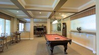 This single-floor amenity space is thoughtfully designed to serve as a premier entertainment and social hub. The standout architectural feature is the elegantly detailed coffered ceiling, framed by crisp white molding and supported by decorative columns, lending a refined, upscale character to the room. Recessed lighting and a classic pendant light fixture illuminate the space warmly throughout.

At the heart of the room sits a full-size pool table with rich, dark wood craftsmanship and a bold red felt surface, making it a natural focal point for recreation. Nearby, a dart board station adds to the game room atmosphere, while a dedicated dining area — complete with a dark wood table, matching chairs, and an entertainment center — provides a comfortable setting for meals or casual gatherings.

The neutral carpet flooring and warm, earthy wall tones create a welcoming ambiance throughout. Large windows along the perimeter allow natural light to filter into the space, enhancing the open, airy feel. A counter-height bar ledge offers additional seating and social space, making this room equally suited for both active play and relaxed conversation. Overall, the space balances classic design with versatile functionality.