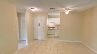 This single-floor space showcases an open-concept layout that seamlessly connects the main living area to the kitchen. The interior is finished with large, light-toned ceramic floor tiles that run continuously throughout, contributing to a clean and cohesive aesthetic. Walls are painted in a soft, warm neutral tone, creating a bright and airy atmosphere.

The kitchen is situated at the rear of the open space and is equipped with white cabinetry offering ample storage, a white electric range, and a sink positioned beneath a window that allows natural light to filter in. The white appliances and cabinetry maintain a consistent, tidy appearance throughout the cooking area.

A ceiling fan with an integrated light fixture is installed in the main living area, providing both ventilation and overhead lighting. An additional flush-mount ceiling light further supplements illumination. A louvered closet door is visible to one side, suggesting convenient built-in storage. The entry door features a mail slot, adding a practical touch. Overall, the space presents a straightforward, functional layout with a light and neutral palette.