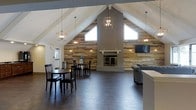 This inviting single-floor gathering space makes a strong first impression with its dramatic vaulted ceilings that rise to a sharp peak, creating an expansive, open atmosphere. The centerpiece of the room is a floor-to-ceiling fireplace framed by a crisp white mantel and surround, set against a striking reclaimed wood accent wall that spans the full width of the space. The warm, rustic texture of the wood paneling adds character and depth, complementing the dark hardwood flooring that runs throughout. Natural light filters in through strategically placed upper-level windows flanking the fireplace, brightening the interior while highlighting the architectural details overhead. A series of geometric lantern-style pendant lights hang from the ceiling, providing both ambient illumination and a contemporary design element. The space is thoughtfully arranged with a casual dining area featuring dark wood tables and chairs on one side, and a relaxed lounge seating area anchored by a gray sofa on the other. A wall-mounted flat-screen television is positioned near the fireplace for added functionality. The overall ambiance strikes a balance between rustic warmth and modern design, making it well-suited for community gatherings, social events, or everyday relaxation.