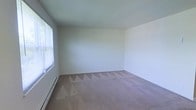 This single-floor space presents a clean, minimalist interior defined by its open layout and neutral aesthetic. The main room is generously sized, offering ample floor space covered in beige wall-to-wall carpeting that adds warmth and comfort underfoot. The walls are finished in a soft, off-white tone that creates a bright and airy atmosphere throughout. A large window fitted with horizontal blinds is positioned along one wall, allowing natural light to filter into the room while offering privacy control. A baseboard heating unit runs along the base of the window wall, providing an efficient and unobtrusive heat source. The ceiling is smooth and painted in a matching light tone, contributing to the overall sense of openness in the room. The space is currently vacant, making it easy to envision a variety of furniture arrangements and uses. The straightforward, no-frills design provides a versatile and functional environment well-suited for residential use. The combination of neutral finishes, natural light, and soft carpeting gives the space a quiet, comfortable character that serves as a solid foundation for personalization.