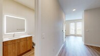 This single-floor space presents a cohesive modern design characterized by neutral wall tones, clean white trim, and wood-style flooring that flows consistently throughout the unit. The open layout connects key living areas, creating a functional and visually unified interior.

The kitchen is well-appointed with a combination of white upper cabinetry and warm wood-toned lower cabinets, complemented by a white tile backsplash and light countertops. Stainless steel appliances, including a refrigerator with ice and water dispenser, dishwasher, range, and over-the-range microwave, provide a complete and practical cooking environment. Natural light enters through a window adjacent to the refrigerator.

The bathroom features a warm wood-toned vanity with ample drawer storage, a white countertop, and a backlit frameless mirror. A full soaking tub with a tiled surround in neutral gray and brushed nickel fixtures completes the space, finished with light wood-style flooring.

The living areas are bright and open, benefiting from multiple windows and glass-paneled exterior doors that allow natural light to fill the space. A hallway connects the bathroom and living areas, and a louvered closet door adds a functional architectural detail throughout.