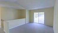 This single-floor space offers a clean and functional layout designed for comfortable everyday living. The main living area is finished with soft, neutral-toned carpet that extends throughout, providing a warm and inviting feel underfoot. The walls are painted in a soft yellow hue, contributing to a light and airy ambiance that makes the space feel open and welcoming.

A standout feature of the living area is the large sliding glass door, which floods the room with natural light and provides direct access to an outdoor balcony. Through the glass, views of surrounding greenery and neighboring structures are visible, enhancing the connection between interior and exterior. The white-railed balcony adds a practical and aesthetic outdoor extension to the living space.

The room features clean white baseboards that contrast neatly against the carpeted floors, and the ceiling is smooth and unadorned, maintaining a simple, understated aesthetic. A partial wall divider adds architectural interest and helps define areas within the open floor plan without fully enclosing the space. Overall, the space presents a straightforward, well-lit interior that is move-in ready and easy to personalize.
