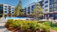Located at 407 Pratt Ave NE in Huntsville, Alabama, this outdoor amenity space is centered around a generously sized resort-style swimming pool that serves as the focal point of a well-appointed courtyard. The pool features a classic curved design with crystal-clear blue water, built-in submerged lounge areas, and easy-access entry points via wide steps and handrails.

The surrounding pool deck is finished in textured concrete and lined with an ample supply of modern chaise lounges and green-accented market umbrellas, providing comfortable spaces for sunbathing and relaxation. A central gathering area includes a circular fire pit set on a patch of artificial turf, offering an inviting social space separate from the pool.

The courtyard is enclosed on multiple sides by a four-story contemporary building featuring a mix of white and warm neutral facades, large windows, and private balconies that overlook the amenity area. Stone-clad base accents, dark window framing, and varied exterior materials give the architecture a modern yet refined character.

Mature trees and manicured landscaping line the perimeter of the courtyard, providing natural greenery and shade throughout the space. The overall ambiance is that of an upscale, resort-inspired outdoor retreat designed for both active recreation and leisurely relaxation.