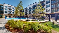Located at 407 Pratt Ave NE in Huntsville, Alabama, this outdoor amenity space is centered around a generously sized resort-style swimming pool that serves as the focal point of a well-appointed courtyard. The pool features a classic curved design with crystal-clear blue water, built-in submerged lounge areas, and easy-access entry points via wide steps and handrails.

The surrounding pool deck is finished in textured concrete and lined with an ample supply of modern chaise lounges and green-accented market umbrellas, providing comfortable spaces for sunbathing and relaxation. A central gathering area includes a circular fire pit set on a patch of artificial turf, offering an inviting social space separate from the pool.

The courtyard is enclosed on multiple sides by a four-story contemporary building featuring a mix of white and warm neutral facades, large windows, and private balconies that overlook the amenity area. Stone-clad base accents, dark window framing, and varied exterior materials give the architecture a modern yet refined character.

Mature trees and manicured landscaping line the perimeter of the courtyard, providing natural greenery and shade throughout the space. The overall ambiance is that of an upscale, resort-inspired outdoor retreat designed for both active recreation and leisurely relaxation.