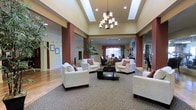 This single-floor community space exudes a warm and inviting ambiance, thoughtfully designed to serve as a social hub for residents and guests alike. The centerpiece of the main lounge is a striking multi-arm chandelier suspended from an elevated tray ceiling, which adds a sense of grandeur to the open-plan layout. Recessed lighting complements the chandelier, ensuring the space is well-lit throughout.

The lounge area is furnished with plush white armchairs and sofas, accented by decorative red and brown throw pillows that echo the rich, earthy tones of the architectural columns. A dark espresso coffee table anchors the seating arrangement atop a patterned area rug, while warm hardwood flooring extends throughout the surrounding areas.

Tall, terracotta-toned accent columns frame the space, creating a natural division between the main lounge and adjacent areas, which appear to include a dining or activity room with dark wood furniture and drapery-framed windows that allow natural light to filter in. Lush indoor greenery adds a refreshing, natural element to the décor.

Additional spaces visible beyond the lounge suggest a fitness area or multipurpose room, making this a versatile, amenity-rich environment designed for comfort and community engagement.