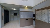 This building lobby serves as a functional and welcoming entry point for residents. The space is designed with practicality in mind, highlighted by a centralized mail and package center that forms the focal point of the area. The mail station features an extensive array of individual resident mailboxes, neatly arranged in a grid formation on a modern, wood-toned panel, providing each resident with secure and organized mail access. Adjacent to the main mailbox unit, a separate parcel locker system accommodates package deliveries, ensuring residents can retrieve their items at their convenience. A built-in outgoing mail slot is integrated into the lower portion of the mail station, adding further functionality to the space. The lobby is finished with light wood-look tile flooring that lends a clean and contemporary aesthetic throughout. Walls are painted in a neutral gray tone, contributing to a calm and orderly atmosphere. Recessed and flush-mounted lighting illuminates the area evenly, while the high ceiling enhances the sense of openness. An access door with a small window is visible toward the rear, connecting the lobby to the broader building interior. Overall, this lobby is thoughtfully designed to meet the everyday needs of building residents efficiently.