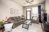 This single-floor unit offers a thoughtfully designed layout that balances modern aesthetics with everyday functionality. The open-concept living area is bright and welcoming, featuring a ceiling fan with integrated lighting, a glass-paneled exterior door that invites natural light, and access to an outdoor balcony. The space flows seamlessly into the kitchen, which is outfitted with dark espresso cabinetry, stainless steel appliances including a refrigerator, range, microwave, and dishwasher, as well as light gray countertops and open floating shelves. A central kitchen island with bar seating provides additional prep and dining space, while warm hardwood-style flooring runs throughout, tying the common areas together cohesively. The kitchen benefits from notably elevated ceilings, adding a sense of openness to the space. The bedroom is comfortably sized and receives generous natural light through a large window with outdoor views of the surrounding area. The bathroom connects to a dedicated laundry area equipped with a stacked washer and dryer, offering added convenience. Overall, the unit presents a clean, contemporary aesthetic with neutral tones, modern finishes, and a practical, well-organized floor plan.