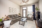 This single-floor unit offers a thoughtfully designed layout that balances modern aesthetics with everyday functionality. The open-concept living area is bright and welcoming, featuring a ceiling fan with integrated lighting, a glass-paneled exterior door that invites natural light, and access to an outdoor balcony. The space flows seamlessly into the kitchen, which is outfitted with dark espresso cabinetry, stainless steel appliances including a refrigerator, range, microwave, and dishwasher, as well as light gray countertops and open floating shelves. A central kitchen island with bar seating provides additional prep and dining space, while warm hardwood-style flooring runs throughout, tying the common areas together cohesively. The kitchen benefits from notably elevated ceilings, adding a sense of openness to the space. The bedroom is comfortably sized and receives generous natural light through a large window with outdoor views of the surrounding area. The bathroom connects to a dedicated laundry area equipped with a stacked washer and dryer, offering added convenience. Overall, the unit presents a clean, contemporary aesthetic with neutral tones, modern finishes, and a practical, well-organized floor plan.