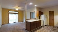 This single-story home offers an open-concept layout that seamlessly connects the kitchen and living areas. The kitchen is equipped with dark wood cabinetry, a double-basin sink set into a light-colored countertop, a white refrigerator, and a dishwasher. Upper wood cabinets provide ample storage, and a pass-through window connects the kitchen to an adjacent space, enhancing the open feel. Recessed lighting illuminates the kitchen area, while a ceiling fan with a light fixture serves the carpeted living room. The living area benefits from abundant natural light through a large sliding glass door, which provides views of an outdoor green space. The kitchen features durable tile flooring, contrasting with the plush carpet of the living room. A structural column adds an architectural element that subtly defines the transition between the two spaces. Overall, the home presents a functional and straightforward layout suited for comfortable everyday living.