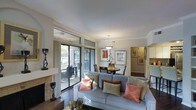 This single-floor residence showcases a thoughtfully designed open-concept layout that seamlessly connects the living, dining, and kitchen areas. The living room serves as the centerpiece of the space, anchored by a classic fireplace with a stone surround and framed by large sliding glass doors that flood the room with natural light and provide direct access to a private outdoor patio. Crown molding runs along the ceiling, adding a refined architectural detail throughout.

The living area is generously proportioned, comfortably accommodating a full-sized sofa adorned with decorative throw pillows in warm coral and neutral tones. Adjacent to the living space, a dedicated dining area features a round table beneath a pendant light fixture, creating an intimate setting for meals. Framed artwork on the neutral-toned walls adds personality and warmth to the space.

A glimpse into the adjoining hallway reveals additional rooms beyond, suggesting a well-organized floor plan. The kitchen, partially visible, appears bright and functional. Dark hardwood-style flooring grounds the space and provides a rich contrast to the light walls and soft furnishings, contributing to the overall warm and inviting ambiance of this well-appointed residence.