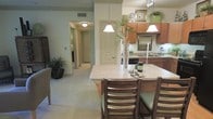 This single-floor residence is designed with an open, flowing layout that seamlessly connects the kitchen, dining, and living areas. The kitchen is a standout feature, outfitted with rich warm-toned wood cabinetry, a smooth light-colored countertop breakfast bar, and a full suite of appliances. Pendant lights hang elegantly above the bar, casting a warm glow over the dining seating — a pair of wooden chairs with green upholstered cushions that add a welcoming touch. Upper cabinets extend to the ceiling, maximizing storage space, while a track lighting system illuminates the work surfaces below. A recessed sink and additional counter space along the back wall make this kitchen both functional and stylish. The adjoining hallway, lit by a flush-mount ceiling fixture, leads to additional rooms and is lined with neutral carpet flooring that continues throughout the living areas. Potted greenery, including tall bamboo-like plants and a lush floor plant, are positioned throughout the space, adding natural warmth and vibrancy. The overall palette of warm beige tones, natural wood finishes, and soft green accents creates a cohesive and inviting ambiance throughout this thoughtfully designed home.