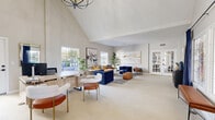 Located at 5242 Edmondson Pike in Nashville, TN, this single-floor space makes a striking first impression with its dramatic vaulted ceilings that draw the eye upward and lend an expansive, airy quality to the interior. The open-plan living room is bathed in natural light, courtesy of multiple large windows dressed with deep navy curtains that add a bold contrast to the otherwise soft, neutral palette of warm whites and light tones.

A standout architectural feature is the floor-to-ceiling whitewashed brick accent wall, which adds texture and character while complementing the contemporary design aesthetic. Light-toned hardwood flooring runs throughout, enhancing the brightness of the space. Black-framed French doors and large glass panel doors provide seamless transitions between areas and connect the interior to the outdoors.

The living area is thoughtfully arranged with a mix of seating, including channeled navy blue armchairs, a mustard-toned sofa, and modern accent chairs in neutral bouclé fabric, all centered around a sleek coffee table. A dedicated workspace with a light wood desk sits near the front windows, making this space as functional as it is inviting. Curated wall art, lush indoor plants, and layered textures complete the polished, welcoming ambiance of this Nashville property.