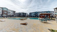 Located at 375 Greenbriar Rd in Fate, TX, this impressive outdoor amenity complex is designed to deliver a resort-style living experience. At its centerpiece sits a generously sized swimming pool surrounded by an expansive stone-paved deck, lined with numerous lounge chairs ideal for relaxing in the Texas sun. A built-in spa or water feature adds an elevated touch to the poolside environment. A covered pavilion with a pitched roof anchors one corner of the deck, providing a shaded gathering space furnished with seating for outdoor dining and socializing. Cornhole boards on the deck further enhance the recreational atmosphere. Adjacent to the pool area, a dedicated pickleball and tennis court offers a vibrant blue-and-green playing surface enclosed by chain-link fencing, with benches positioned courtside for spectators. Well-maintained landscaping with manicured lawn areas and young plantings borders the courts, adding greenery to the setting. The surrounding multi-story residential buildings, featuring a contemporary mix of brick, board-and-batten siding, and private balconies, frame the amenity space and contribute to its cohesive community aesthetic. Winding pathways connect the various outdoor zones, creating a seamless and accessible layout throughout this well-appointed outdoor community hub.