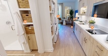 This thoughtfully designed 290-square-foot, one-bedroom, one-bathroom apartment maximizes every inch of its single-floor layout. The galley-style kitchen is a standout feature, outfitted with light wood cabinetry, white quartz countertops, a stainless steel sink, built-in dishwasher, induction cooktop, microwave, and a full-size bottom-freezer refrigerator. Under-cabinet lighting and open display shelving add both function and style, while light wood-look flooring runs seamlessly throughout the unit. The 160-square-foot primary bedroom offers a calm, comfortable retreat, featuring a large window with views of surrounding greenery and architecture, providing ample natural light. The room comfortably accommodates a queen bed and includes closet storage and a wall-mounted television area. The bathroom is finished in a crisp, all-white palette with light wood floors, a glass-enclosed shower, open built-in shelving for storage, and a clean, minimal aesthetic. The overall design language of the apartment is modern and cohesive, combining warm wood tones, white surfaces, and neutral accents to create a bright and functional living space well-suited for single occupancy or as a compact urban dwelling.