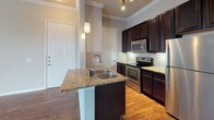 Located at 1316 Town Center Dr in Pflugerville, Texas, this single-floor unit offers a well-designed layout that balances functionality with modern finishes. Rich, dark hardwood-style flooring extends throughout the living areas and bedrooms, providing a warm and cohesive aesthetic anchored by crisp white crown molding and neutral gray walls.

The kitchen is a standout feature, showcasing espresso-toned cabinetry that reaches toward the ceiling, granite countertops, and a central island with a double stainless steel sink. Pendant lighting above the island adds a refined touch, while a full suite of stainless steel appliances — including an electric range, over-range microwave, and refrigerator — completes the space. A classic subway tile backsplash and tile accent wall add additional texture and character.

The open living and dining area flows naturally from the kitchen, benefiting from multiple doorways that connect to the bedrooms and additional rooms. The bedrooms are well-proportioned, each featuring large windows fitted with horizontal blinds that invite natural light while maintaining privacy. Crown molding carries through these rooms, reinforcing the consistent design language found throughout the unit.

Overall, the space presents a clean, move-in-ready environment with thoughtful finishes and a practical single-level floor plan suited for comfortable everyday living.