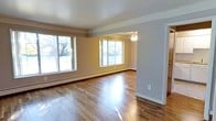 This single-floor residence offers a clean, functional layout with a welcoming ambiance throughout. The main living area is bright and spacious, featuring rich hardwood flooring that reflects natural light pouring in through large, multi-pane windows. Baseboard heating lines the walls, and a ceiling fan with a built-in light fixture provides year-round comfort. The open feel is enhanced by neutral-toned walls and an unobstructed flow between the living and adjoining dining or secondary living area, which shares the same hardwood floors, ceiling fan, and generous window views of the surrounding greenery.

The kitchen is equipped with white cabinetry offering ample upper and lower storage, a stainless steel sink, laminate countertops, and a white gas range with an overhead exhaust hood. The tile flooring provides a practical and easy-to-maintain surface in this well-organized cooking space.

The bathroom features a full-sized soaking tub with a tiled surround accented by a classic border pattern in muted lavender and cream tones. A separate shower area, a vanity with an integrated sink, and mosaic tile flooring complete the space. Overall, this residence presents a straightforward and comfortable living environment with functional finishes throughout.