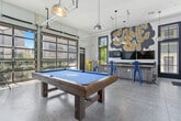 Located at 2315 South 11th Avenue in Bozeman, Montana, this single-floor game room delivers a lively, mountain-inspired atmosphere with a distinctly urban edge. The open-plan layout makes excellent use of its generous footprint, with polished concrete floors and elevated ceilings that amplify the sense of space throughout.

Floor-to-ceiling glass garage doors line one wall, flooding the interior with abundant natural light and offering views of the surrounding neighborhood. Industrial-style pendant lights hang from exposed ceiling infrastructure, reinforcing the room's modern, utilitarian aesthetic.

A central pool table with a bold blue felt surface serves as the main focal point, complemented by a rustic wood-paneled bar counter with a dark granite top and colorful metal barstools in yellow and blue. A dedicated kitchenette area features dark wood cabinetry, a granite countertop, a mounted TV, and a striking hand-lettered "Game On" mural that commands attention.

A mountain-themed art installation with a "Ski Repair" sign anchors one corner, nodding to Bozeman's outdoor recreation culture and adding a locally inspired decorative element. The space also includes additional seating along the glass doors, creating an ideal area for socializing. Overall, this game room combines playful design, functional amenities, and regional character into a cohesive and energetic communal space.