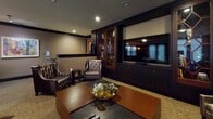 Located at 1200 N Garfield Street in Arlington, Virginia, this elegantly appointed amenity lounge offers a refined, multi-functional gathering space thoughtfully designed for socializing and relaxation. The single-floor layout is anchored by a striking curved bar with a dark granite countertop, sleek black cabinetry, and pendant lighting featuring warm amber glass fixtures that cast an inviting glow. High-top bar seating lines the bar area, while a separate dining zone with dark wood tables and upholstered chairs provides a more formal setting. A vibrant large-scale abstract painting adds a bold focal point to the main lounge area, which features a sculptural curved sofa, tufted leather armchairs, and a rich wood coffee table. Built-in dark wood shelving displays curated books and décor, complementing the space's sophisticated palette of deep browns, warm taupes, and charcoal tones. A dedicated media lounge area includes a large flat-screen television mounted within custom dark wood cabinetry, alongside a glass-door wine display cabinet with decorative diamond-patterned woodwork. Patterned carpeting runs throughout, and track lighting along the ceiling provides adjustable ambient illumination. Large windows with horizontal blinds allow natural light to filter into the space, enhancing the upscale yet comfortable atmosphere throughout.