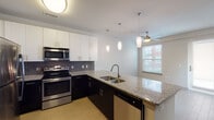 This thoughtfully designed single-floor residence offers a cohesive blend of modern finishes and practical functionality throughout. The heart of the home is the open-concept kitchen, which features striking two-tone cabinetry — crisp white upper cabinets paired with dark lower cabinets — set against a mosaic tile backsplash. Granite countertops extend across the workspace and wrap around a large peninsula with an undermount sink, providing ample prep and entertaining space. A full suite of stainless steel appliances, including a range, microwave, refrigerator, and dishwasher, completes the kitchen. Pendant lighting hangs above the peninsula, while a ceiling fan positioned in the adjacent living area ensures year-round comfort. Natural light filters in through a window overlooking the exterior. The bedroom is a generous, carpeted space with neutral walls that offer a calm, versatile backdrop. A dedicated walk-in closet provides substantial storage with wrap-around shelving and hanging rails. The bathroom is finished with the same granite countertop seen in the kitchen, paired with dark cabinetry, an undermount sink, and a large framed mirror with overhead lighting. In-unit laundry is conveniently tucked into a dedicated closet, housing a front-load washer and dryer. The overall design is clean, contemporary, and move-in ready.