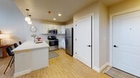 This thoughtfully designed single-floor residence combines industrial character with modern comfort. The living area commands attention with dramatically high ceilings and a bank of glass block windows that flood the space with diffused natural light. A sunburst mirror anchors the neutral-toned walls, while light hardwood flooring, a bold geometric area rug, and mid-century inspired furnishings contribute to a warm, eclectic ambiance. A wall-mounted mini-split unit ensures year-round climate comfort.

The kitchen is both functional and stylish, featuring white shaker cabinetry, gleaming white countertops, and a full suite of stainless steel appliances including a range, microwave, and refrigerator. A peninsula with seating for three, illuminated by globe pendant lights, creates an inviting space for casual dining and entertaining.

The bedroom continues the airy atmosphere with the same soaring ceiling height and glass block windows positioned high on the wall, allowing for privacy while still drawing in natural light. Light hardwood flooring and ample wall space make this a restful retreat.

The full bathroom is clean and efficiently laid out, with a tub-shower combination, a dark-finished vanity with a white countertop, and light tile flooring accented by a vibrant runner rug. The overall design of this space balances openness and intimacy with a cohesive, contemporary aesthetic throughout.