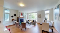 This well-appointed two-floor space combines modern comfort with warm, inviting design throughout. The main living area immediately impresses with its gleaming hardwood floors, crisp white walls, and an abundance of natural light streaming in through large, floor-to-ceiling windows that frame verdant outdoor views. A standout feature of the living room is the classic tile-surround fireplace with a warm wood mantle, adding a cozy focal point to the space. The open layout seamlessly connects the living and dining areas, creating an ideal environment for both relaxing and entertaining. Contemporary furnishings complement the clean architectural lines, while tasteful wall art and decorative accents contribute to the polished, curated aesthetic. The generous window placement ensures that natural light fills the space throughout the day, enhancing the airy and open ambiance. With two floors, the layout offers ample room for a variety of living arrangements, providing both communal spaces for gathering and private areas for retreat. The overall design strikes a balance between functional living and stylish comfort, making this a versatile and welcoming space.
