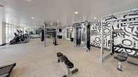 Located at 5555 E Mockingbird Ln in Dallas, Texas, this athletic club fitness center is a comprehensively outfitted facility designed to support a wide range of workout routines. The space is organized into distinct functional zones, offering both cardio and strength training areas within a single open floor plan. The cardio section features a row of Matrix treadmills and elliptical machines positioned alongside expansive floor-to-ceiling windows, providing unobstructed views of the outdoor pool and surrounding courtyard. Natural light floods this area, creating an inviting and energizing environment for workouts. The strength training zone is equally impressive, with a cable machine, adjustable weight bench, pull-up and dip rig, and a heavy punching bag. A dedicated free weights area houses a comprehensive rack of kettlebells and medicine balls, along with TRX suspension trainers and resistance equipment. The room's design is accentuated by a bold black-and-white graphic mural that covers an entire wall, adding a dynamic and modern aesthetic to the space. Full-length mirrors reflect light and give the room an expansive feel. Neutral carpet flooring, recessed lighting, and clean white ceilings complete the polished, contemporary interior, making this facility a standout amenity.