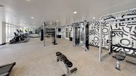 Located at 5555 E Mockingbird Ln in Dallas, Texas, this athletic club fitness center is a comprehensively outfitted facility designed to support a wide range of workout routines. The space is organized into distinct functional zones, offering both cardio and strength training areas within a single open floor plan. The cardio section features a row of Matrix treadmills and elliptical machines positioned alongside expansive floor-to-ceiling windows, providing unobstructed views of the outdoor pool and surrounding courtyard. Natural light floods this area, creating an inviting and energizing environment for workouts. The strength training zone is equally impressive, with a cable machine, adjustable weight bench, pull-up and dip rig, and a heavy punching bag. A dedicated free weights area houses a comprehensive rack of kettlebells and medicine balls, along with TRX suspension trainers and resistance equipment. The room's design is accentuated by a bold black-and-white graphic mural that covers an entire wall, adding a dynamic and modern aesthetic to the space. Full-length mirrors reflect light and give the room an expansive feel. Neutral carpet flooring, recessed lighting, and clean white ceilings complete the polished, contemporary interior, making this facility a standout amenity.