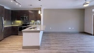 Located at Aura 360 Flats in Grand Prairie, Texas, this single-story unit features a thoughtfully designed open-concept layout connecting the kitchen and living areas. The kitchen is outfitted with dark espresso cabinetry, granite countertops, a green glass tile backsplash, and a full suite of stainless steel appliances, including a refrigerator, electric range, and built-in microwave. A peninsula with an undermount sink and pendant lighting above serves as a natural divider between the kitchen and the open living area, which continues the light wood-style vinyl plank flooring found throughout the main areas. The entryway, visible from the kitchen, offers a practical transition into the unit. The bedroom features plush carpeting, a ceiling fan, and a window with blinds that allows natural light to filter in. The bathroom presents a spacious, well-appointed design with dark cabinetry, granite countertops, an undermount sink, a soaking tub with tiled surround, and wood-style flooring consistent with the rest of the unit. A walk-in closet is accessible directly from the bathroom, adding functional storage. The overall aesthetic is clean and contemporary, with neutral tones and modern finishes throughout.