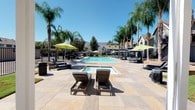 This inviting outdoor amenity space centers around a generously sized swimming pool with crystal-clear blue water, complemented by a separate spa for relaxation. The expansive stone-tiled sundeck provides ample room for lounging, furnished with cushioned chaise lounges and shade umbrellas in a vibrant lime green, creating a lively, resort-inspired atmosphere. Towering palm trees and mature shade trees are thoughtfully positioned throughout the area, lending a tropical ambiance and natural privacy. Multiple covered pergola-style cabanas run along the poolside, each equipped with slatted benches and tables, offering shaded gathering spots ideal for outdoor dining or socializing. The entire pool area is enclosed by a sleek black metal security fence, ensuring a safe and well-defined environment. In the background, the surrounding residential buildings and mountain ridgeline are visible under a clear blue sky, reinforcing the Southern California setting. Decorative rock landscaping fills garden beds along the perimeter, adding low-maintenance greenery that complements the overall design. The combination of sun-drenched open deck space and shaded retreat areas makes this outdoor space well-suited for both active recreation and relaxed lounging throughout the day.