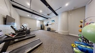 This fitness center combines historic architectural character with modern exercise amenities. The space is defined by its white-painted brick walls, striking dark exposed ceiling beams, and a notable cylindrical brick column that serves as a distinctive structural focal point. Recessed lighting and a ceiling fan are mounted above, keeping the space bright and well-ventilated throughout.

The floor is covered in durable patterned carpet tile suited for a high-traffic workout environment. Natural light enters through arched and rectangular windows, adding warmth to the otherwise industrial aesthetic. A wall-mounted flat-screen television is positioned for easy viewing during cardio sessions, and a mini-split HVAC unit ensures climate control year-round.

Cardio equipment includes treadmills, an elliptical, and a stationary bike, all lined along one wall. A multi-tiered dumbbell rack stocked with a full range of free weights sits alongside a flat workout bench, while a cable machine provides resistance training options. A medicine ball rack rounds out the strength training offerings.

A frosted-glass door leads to an adjoining room, and a sanitation station with a waste bin is conveniently placed near the entrance. The overall ambiance is functional yet full of character, blending the charm of historic architecture with the practical demands of a modern fitness facility.