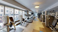 This fitness center is a spacious, well-appointed facility designed to accommodate a wide range of workout routines. The layout is thoughtfully organized, with distinct zones dedicated to cardiovascular equipment, strength training machines, and free weights. Along one side of the room, floor-to-ceiling windows allow abundant natural light to pour in while offering views of the surrounding urban streetscape, creating an energizing atmosphere for workouts.

The cardio zone features a row of treadmills, elliptical trainers, and stationary bikes, all positioned to take advantage of the natural light and outdoor views. Adjacent to this area, a collection of multi-station cable machines and selectorized strength equipment provides versatile options for resistance training. A dedicated free weights section includes a fully stocked dumbbell rack, complemented by an adjustable workout bench.

Large mirrors line one wall, visually expanding the space and allowing users to monitor their form. The flooring combines smooth hard surfaces with rubber mat insets beneath equipment for added traction and protection. Overhead, pendant light fixtures supplement the natural light. The color palette of neutral tones with accents of navy blue gives the space a clean, modern aesthetic.