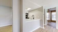 This thoughtfully updated single-floor residence showcases a clean, contemporary aesthetic throughout. The kitchen serves as a functional centerpiece, featuring crisp white shaker cabinets, striking marble-look quartz countertops, a bold black hexagon tile backsplash, and a full suite of stainless steel appliances. A peninsula with a matte black faucet and integrated sink opens toward the living area, promoting a connected, open-concept feel. Light-toned luxury vinyl plank flooring flows consistently throughout the space, complementing the neutral warm-gray wall palette. The living area is generously proportioned with soaring vaulted ceilings and a ceiling fan, and sliding glass doors with vertical blinds open to a view of an outdoor pool area, inviting abundant natural light. The bedroom with walk-in closet access offers a private, functional retreat with its own natural light via a window with blinds. The bathroom is finished with a floating white vanity, matte black hardware and fixtures, a backlit mirror, and light wood-tone flooring that ties seamlessly to the rest of the home. Overall, the space presents a cohesive modern design with carefully selected fixtures and finishes throughout.