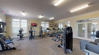 Located at 56 Bent Branch Loop Rd in Clayton, NC, this well-appointed fitness center is designed to support a full range of workout routines within a single, open-plan floor. The space features warm yellow-toned walls accented by crisp white crown molding, complemented by durable patterned carpet flooring throughout. Fluorescent overhead lighting ensures the entire facility is evenly and brightly illuminated, while multiple windows fitted with blinds allow for natural light and views of the surrounding area.

The cardio section is equipped with multiple treadmills and elliptical cross-trainers, each paired with wall-mounted flat-screen televisions to keep users engaged during their workouts. Large wall mirrors line portions of the space, visually expanding the room and allowing for form monitoring during exercise.

The strength training area offers a diverse selection of cable machines, multi-station weight systems, and selectorized resistance equipment, providing options for targeting a wide variety of muscle groups. Additional accessories such as medicine balls and TRX suspension trainers are also present, supporting functional and bodyweight training.

Glass-paned doors provide access to the facility, contributing to an open and inviting atmosphere. Overall, the fitness center is thoughtfully organized to maximize usable floor space while offering a comprehensive and comfortable environment for exercise.