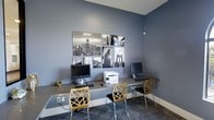 This business center offers a well-appointed and functional workspace designed to support productivity and convenience. The room is finished with cool blue-gray walls that lend a calm, professional atmosphere, complemented by patterned carpet flooring that adds texture underfoot. A built-in countertop desk runs along the main wall, providing ample surface area for multiple users working simultaneously. The workstations are equipped with desktop computers and a shared multifunction printer, making the space fully ready for everyday business tasks. Stylish geometric wood-and-metal chairs offer comfortable seating at each station, blending modern design with practicality. Above the desk, a curated gallery wall featuring a collection of black-and-white cityscape photographs — including iconic views of bridges and skylines — adds visual interest and an urban sophistication to the space. Integrated power outlets along the desk ensure easy access for additional devices. The overall ambiance is sleek and contemporary, striking a balance between a focused work environment and thoughtfully chosen design elements that make the space both inviting and professional.
