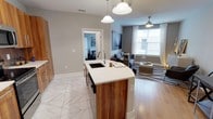 This single-floor residence offers a thoughtfully designed open-concept layout that blends functionality with modern style. The kitchen serves as the centerpiece of the space, anchored by a striking island featuring rich walnut wood cabinetry, a crisp white quartz countertop, and an integrated sink beneath elegant pendant lighting. The kitchen flows naturally into the living area, which is finished with warm hardwood-style flooring and benefits from generous natural light streaming through large windows dressed with sheer curtains. A ceiling fan adds both a practical and aesthetic touch to the living space. The transition between the kitchen and living areas is defined by a shift from large-format marble-look tile flooring to the warm wood tones of the main living space, creating a subtle yet effective delineation of zones. Neutral gray walls provide a versatile backdrop throughout, complementing the contemporary furnishings and décor. A glimpse into an adjacent bedroom reveals a continuation of the clean, modern aesthetic. The overall ambiance is bright, airy, and welcoming, with a cohesive design language that ties every area of the home together seamlessly.