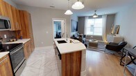 This single-floor residence offers a thoughtfully designed open-concept layout that blends functionality with modern style. The kitchen serves as the centerpiece of the space, anchored by a striking island featuring rich walnut wood cabinetry, a crisp white quartz countertop, and an integrated sink beneath elegant pendant lighting. The kitchen flows naturally into the living area, which is finished with warm hardwood-style flooring and benefits from generous natural light streaming through large windows dressed with sheer curtains. A ceiling fan adds both a practical and aesthetic touch to the living space. The transition between the kitchen and living areas is defined by a shift from large-format marble-look tile flooring to the warm wood tones of the main living space, creating a subtle yet effective delineation of zones. Neutral gray walls provide a versatile backdrop throughout, complementing the contemporary furnishings and décor. A glimpse into an adjacent bedroom reveals a continuation of the clean, modern aesthetic. The overall ambiance is bright, airy, and welcoming, with a cohesive design language that ties every area of the home together seamlessly.