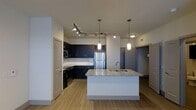 Located in Carrollton, TX, this single-floor residence showcases a thoughtfully designed open-concept layout that blends modern aesthetics with functional living. The kitchen serves as the heart of the space, anchored by a large central island topped with granite countertops and an undermount sink. Dark, rich cabinetry provides striking contrast against the light-toned walls and wood-plank flooring that flows throughout the space. Stainless steel appliances, including a refrigerator, microwave, and range, add a sleek, contemporary finish to the kitchen. Overhead, a combination of track lighting and elegant pendant lights illuminate the cooking and dining areas with warm, layered light. The open floor plan creates a seamless connection between the kitchen and the surrounding living areas, enhancing the sense of space and encouraging a natural flow from room to room. Doors visible throughout suggest well-defined private areas beyond the main living space. The clean lines, neutral color palette, and modern fixtures come together to create an inviting and polished interior well-suited for both everyday living and entertaining.