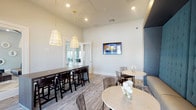 This well-appointed single-floor space functions as a contemporary lounge and social gathering area, thoughtfully designed to accommodate a variety of seating arrangements and uses. The layout is organized around two distinct zones: a communal bar-height table paired with dark wood stools positioned beneath a pair of elegant cone-shaped pendant lights with brass accents, and a relaxed lounge area featuring round marble-topped bistro tables with gold bases and transparent acrylic chairs.

The standout architectural feature is a floor-to-ceiling tufted banquette finished in deep teal velvet, framed by a warm gold-trimmed accent wall. This built-in seating element lends the space a sophisticated, upscale character. A wall-mounted flat-screen television is integrated into the main wall, adding a functional entertainment element to the lounge atmosphere.

Neutral-toned patterned carpet covers the floor throughout, complementing the crisp white walls and light-colored trim. French doors along one wall introduce generous natural light and provide a visual connection to an adjacent outdoor area. Glass-paneled sliding doors on the opposite side open to a neighboring lounge space, enhancing the sense of openness and flow. The overall ambiance is modern and inviting, balancing comfort with refined design.