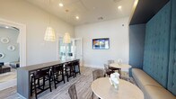 This well-appointed single-floor space functions as a contemporary lounge and social gathering area, thoughtfully designed to accommodate a variety of seating arrangements and uses. The layout is organized around two distinct zones: a communal bar-height table paired with dark wood stools positioned beneath a pair of elegant cone-shaped pendant lights with brass accents, and a relaxed lounge area featuring round marble-topped bistro tables with gold bases and transparent acrylic chairs.

The standout architectural feature is a floor-to-ceiling tufted banquette finished in deep teal velvet, framed by a warm gold-trimmed accent wall. This built-in seating element lends the space a sophisticated, upscale character. A wall-mounted flat-screen television is integrated into the main wall, adding a functional entertainment element to the lounge atmosphere.

Neutral-toned patterned carpet covers the floor throughout, complementing the crisp white walls and light-colored trim. French doors along one wall introduce generous natural light and provide a visual connection to an adjacent outdoor area. Glass-paneled sliding doors on the opposite side open to a neighboring lounge space, enhancing the sense of openness and flow. The overall ambiance is modern and inviting, balancing comfort with refined design.
