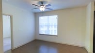 This single-floor space presents a simple, functional layout with a neutral, welcoming ambiance. The room showcases smooth hardwood-style flooring in a warm, dark tone that contrasts cleanly with the light beige walls, creating a balanced and versatile interior palette. A ceiling fan with an integrated light fixture is centrally mounted, providing both air circulation and overhead illumination throughout the space.

A large window fitted with horizontal blinds serves as the room's primary source of natural light, filling the interior with brightness while still offering privacy control. The window's placement enhances the open, airy feel of the room. Clean white baseboards run along the perimeter, adding a finished, polished look to the overall aesthetic.

The walls are smooth and unadorned, offering flexibility for personalization, while multiple electrical outlets are positioned conveniently at various points around the room. The overall condition of the space appears well-maintained and move-in ready. The minimalist design allows the room to function as a living area, bedroom, or multipurpose space depending on the occupant's needs.