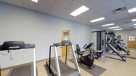 This single-floor fitness room is thoughtfully outfitted with a selection of professional-grade Precor exercise equipment, making it a capable space for both cardiovascular and strength training workouts. The room features multiple treadmills and a recumbent stationary bike for cardio exercise, complemented by a multi-station cable strength machine that supports a wide range of resistance training exercises. Equipment is positioned on protective rubber floor mats, preserving the durable, light-toned flooring beneath. The room's design is clean and utilitarian, with soft blue-gray walls that create a calm workout atmosphere. A full-length wall mirror on one side enhances the sense of space and allows users to monitor their form during exercise. The drop ceiling is fitted with recessed panel lighting that provides bright, even illumination throughout the room. A solid wood-framed door offers a clearly marked entry and exit point. Overall, this fitness room delivers a practical and well-maintained environment suited for regular physical activity, with quality equipment arranged efficiently to maximize usable floor space.