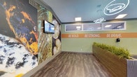 This energetic single-floor commercial space is thoughtfully designed to motivate and engage visitors from the moment they enter. The interior features a striking large-scale nature-inspired wall mural with rich tones of orange, green, and earth hues, creating a vivid focal point that draws the eye immediately. A wall-mounted flat-screen display is integrated seamlessly into this feature wall, adding a functional media element to the artistic backdrop.

The space is anchored by a bold motivational statement — "BE BOLD. BE STRONG." — displayed prominently on a green accent wall with an orange banner, reinforcing a theme of empowerment and vitality. A large planter box filled with ornamental grasses adds a touch of natural warmth and texture to the space.

Overhead, modern ring-shaped pendant lighting fixtures complement the recessed ceiling panels, providing bright, even illumination throughout. The flooring consists of durable wood-look planks in a warm medium tone, offering both practicality and visual appeal. Interior windows allow views into adjacent areas, enhancing the open and connected feel of the layout. The overall ambiance is dynamic, clean, and purpose-driven, making this space well-suited for a fitness, wellness, or youth-focused facility.