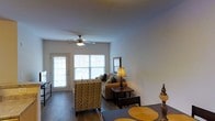 This single-floor residence offers a well-designed open-concept layout that seamlessly connects the living and dining areas. The living room is bright and inviting, benefiting from generous natural light that streams in through large windows and a glass-paneled door, creating an airy atmosphere throughout the space. A ceiling fan with a built-in light fixture is centrally positioned overhead, contributing to both comfort and functionality.

The flooring throughout features hardwood-style planks in a warm, medium-toned finish that adds a clean, cohesive look underfoot. The living area is spacious enough to accommodate a comfortable seating arrangement, including a patterned accent chair and a neutral-toned sofa, complemented by dark wood side tables and a table lamp that adds a warm ambiance.

Adjacent to the living area, the dining space is defined by a dark dining table set with decorative placemats and accents, providing a practical yet stylish area for meals. The walls are finished in a soft, neutral tone that enhances the brightness of the space and provides a versatile backdrop. Overall, this single-floor unit offers a functional, comfortable living environment with a cohesive design aesthetic and ample natural light.