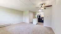 This single-floor unit offers a practical and functional layout with a seamless flow between the living and kitchen areas. The spacious living room is finished with plush carpeting and neutral light green walls, creating a calm and welcoming atmosphere. A ceiling fan with integrated lighting serves as a central fixture, while baseboard heating runs along the perimeter walls for efficient climate control. A wall-mounted air conditioning unit and a large picture window allow natural light to fill the room while offering views of the surrounding greenery outside.

The kitchen is positioned adjacent to the living area, delineated by a peninsula with a light wood-toned countertop that provides additional prep space. White cabinetry lines the walls, complemented by dark granite-style countertops, wood-look flooring, and black appliances including a refrigerator, electric range with overhead ventilation, and a stainless steel sink.

The bedroom features soft carpeting, neutral walls, and a full-width closet with a hanging rod, providing ample storage. The bathroom is finished with wood-look tile flooring, a white vanity with dark countertop, a mirrored medicine cabinet, and a tub-shower combination with a decorative curtain.