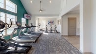 Located at 850 Wilkinson Trce in Bowling Green, Kentucky, this fitness center offers a comprehensive, single-floor exercise facility designed to support a wide range of workout routines. The space features soaring vaulted ceilings and an abundance of natural light streaming through large multi-pane windows, creating an open and energizing atmosphere. A ceiling fan provides added comfort throughout the main workout area.

The cardio zone is prominently positioned along the windowed wall, showcasing a row of treadmills and an elliptical trainer, all with views overlooking an outdoor pool area. Wall-mounted flat-screen televisions are conveniently placed throughout for entertainment during workouts. Adjacent to the cardio section, a dedicated cycling area houses multiple stationary spin bikes on durable carpet tile flooring.

A separate strength and free-weight zone offers a full rack of dumbbells, a weight bench, and additional cable equipment, set on hardwood-style flooring for easy movement. A well-organized accessory station provides resistance bands, medicine balls, and exercise balls in multiple sizes, catering to functional and flexibility training.

The facility features a clean, modern aesthetic with crisp white walls accented by a bold teal feature wall, recessed lighting, and a neutral flooring palette. The thoughtful layout ensures clear zones for various exercise types, making this fitness center a well-rounded and functional amenity.