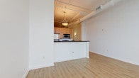 This single-floor loft-style unit showcases industrial-inspired design elements paired with clean, modern finishes throughout. The open living area is anchored by light hardwood flooring that extends seamlessly through the space, while exposed ductwork and structural beams along the ceiling add an authentic urban character. Crisp white walls keep the interiors feeling bright and airy.

The kitchen is positioned behind a peninsula-style breakfast bar topped with a dark countertop, providing both a natural divider from the living area and additional seating space. Inside the kitchen, warm-toned maple cabinetry lines the walls, complemented by a full suite of stainless steel appliances including a refrigerator, electric range, built-in microwave, and dishwasher. Light tile flooring distinguishes the cooking area from the rest of the unit.

The bedroom is a standout feature, offering an expansive wall of large sliding windows that frame sweeping city views, including a prominent clock tower on the skyline. Painted brick below the windows and light hardwood floors lend the room a warm, loft-inspired feel with abundant natural light.

The bathroom is finished in neutral tile and features a pedestal sink, mirrored medicine cabinet, toilet, and a full tub-shower combination, offering a clean and functional layout.