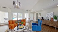 This single-floor community clubhouse is designed with a modern, energetic aesthetic that immediately draws the eye. The main lounge area features soaring vaulted ceilings and rich hardwood-style flooring, creating an open and airy atmosphere. Large glass doors and expansive windows flood the space with natural light while offering direct views of the outdoor pool area, seamlessly connecting the interior to the exterior amenities. A striking geometric pendant chandelier serves as a focal point overhead, complementing the bold interior design choices throughout.

The lounge is furnished with vibrant cobalt blue and tangerine orange velvet seating arranged around a round white coffee table atop a graphic patterned area rug, delivering a lively, contemporary feel. A wooden credenza along one wall provides additional storage and display space, styled with decorative accents and a coffee station. Wall-mounted flat-screen televisions are positioned for easy viewing from multiple seating areas, and a bar-height counter with stools offers a casual gathering spot near the windows.

A dedicated business center off the main lounge provides a focused workspace, featuring a long quartz countertop desk, dual computer workstations, and pendant lighting framed by textured wood-slat and dimensional white wall panels, adding warmth and visual interest to the functional space.