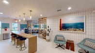 The lounge at 5200 Croyden Ave in Kalamazoo, Michigan is a thoughtfully designed single-floor communal space that blends mid-century modern aesthetics with a warm, inviting atmosphere. The interior is defined by a bold two-tone color palette, featuring teal accent bands along the walls, pops of chartreuse yellow, and crisp white surfaces that together create a lively yet cohesive environment.

The space is organized into several distinct functional zones, elegantly separated by striking slatted wood room dividers that allow light and sightlines to flow freely while providing a sense of structure. A casual dining and social area features a long wooden bar-height table paired with upholstered stools, illuminated by a row of warm brass pendant lights with glass shades. Nearby, a snack and beverage station includes vending machines, a coffee setup, and open shelving for added convenience.

The lounge area offers plush, eclectic seating arrangements including a sectional sofa, a woven rattan chair, and a leather accent chair, all centered around a wall-mounted flat-screen television flanked by modern sconce lighting. Large-format tile flooring transitions to soft carpet in the lounge zone, enhancing comfort underfoot.

A charming built-in nook tucked into a hallway, accented with a chartreuse wall and a warm Edison bulb pendant, provides an intimate retreat. Coastal artwork and lush indoor plants complete the inviting, resort-inspired ambiance throughout.