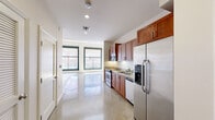 This single-floor unit offers a thoughtfully designed layout that blends modern finishes with functional living spaces. The kitchen serves as a central highlight, featuring rich warm-toned wood cabinetry, granite countertops, and a full suite of stainless steel appliances including a range, microwave, dishwasher, and refrigerator with ice and water dispenser. Polished concrete floors extend throughout the open-plan living and kitchen areas, lending an industrial-chic aesthetic that is both durable and visually cohesive. Recessed lighting and pendant fixtures provide layered illumination throughout the space. The entry foyer is defined by a distinctive solid wood door with a transom window, flanked by interior doors offering closet and utility access. The living area transitions into a sun-filled space distinguished by large, black-framed windows with roll-down shades, offering expansive urban views and abundant natural light. A unique decorative mosaic tile border accents the floor near the windows, adding architectural character. Exposed ceiling beams further enhance the industrial aesthetic of the living zone. The bathroom is finished with classic white subway tile surrounding a full soaking tub and shower combination, complemented by a granite-topped vanity and tile flooring, creating a clean and timeless look.